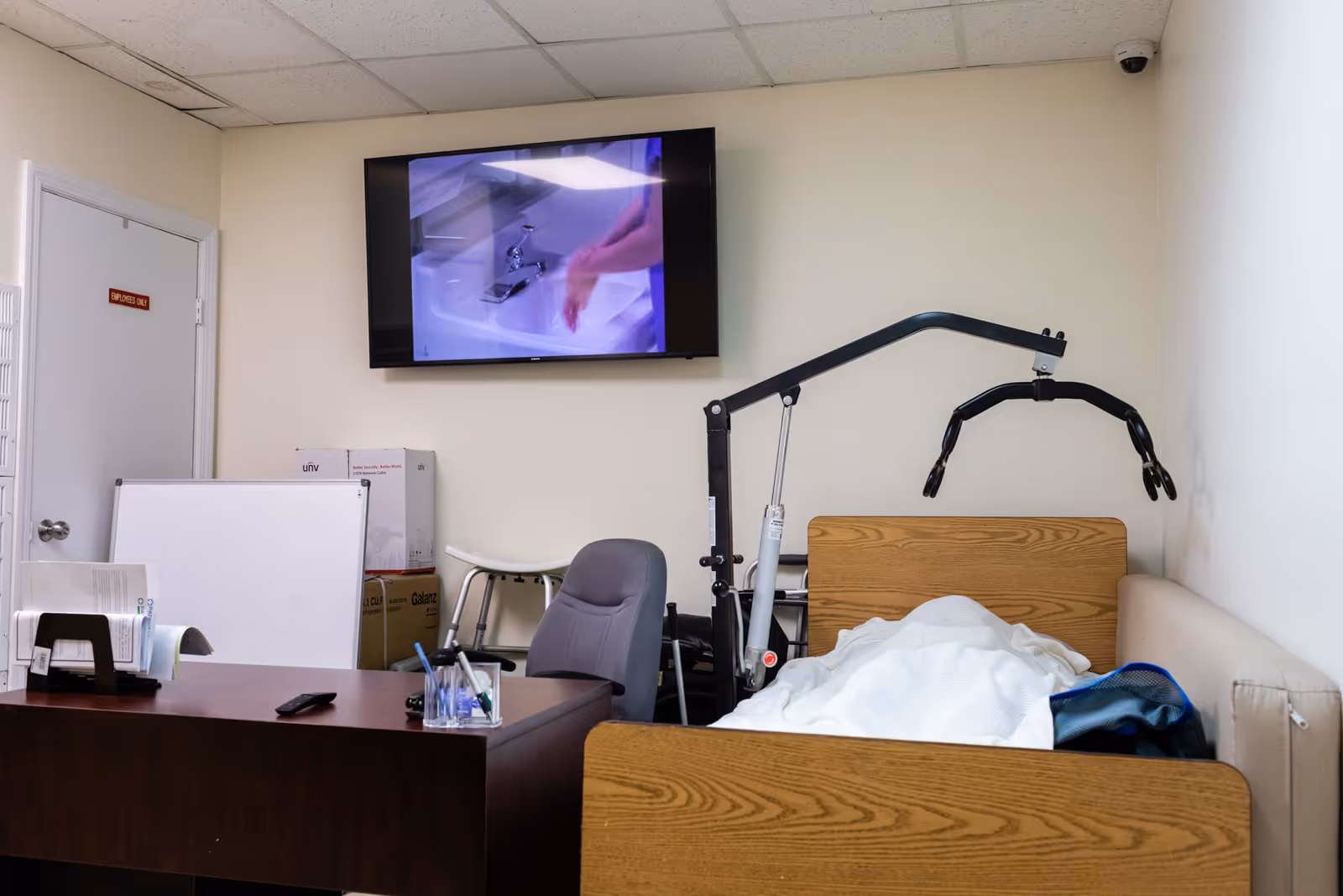 Interior patient room with bed, ceiling lift, desk, chair, and wall-mounted TV.