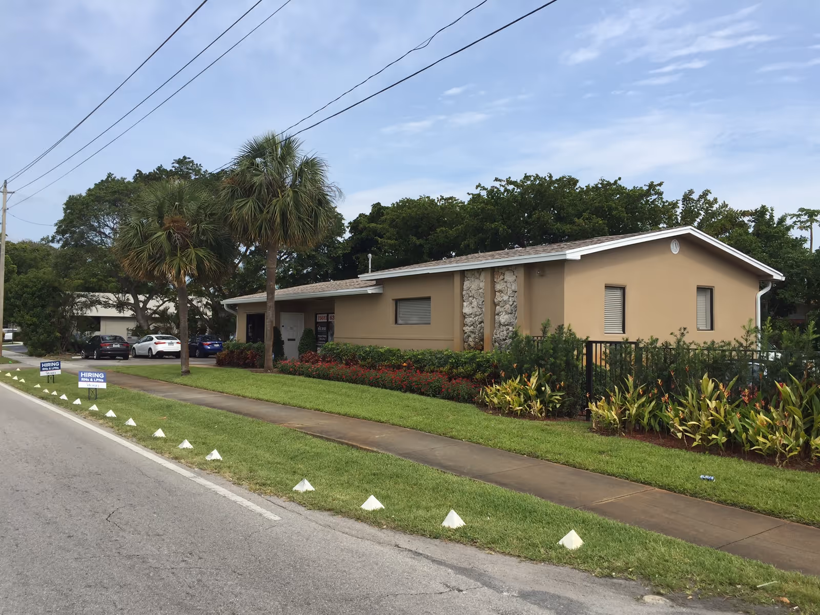Single-story beige facility with stone columns, palm trees, and a manicured lawn along the street
