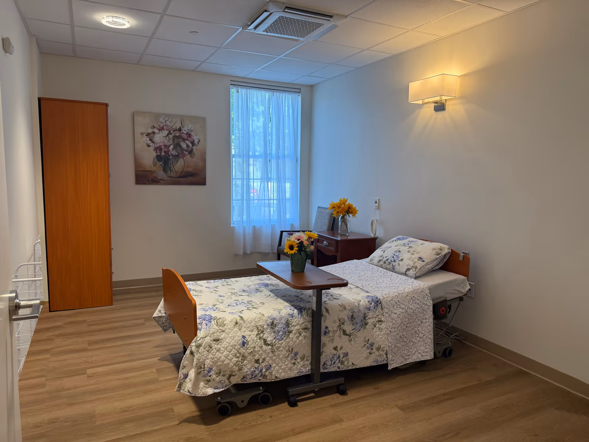 A tidy bedroom with a floral quilted bed, wardrobe, nightstand, and window in soft lighting.