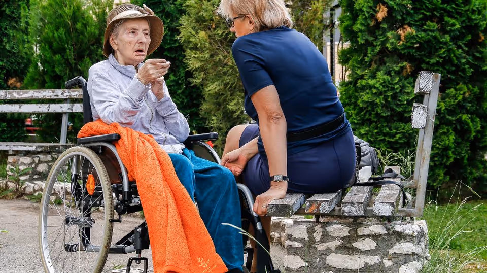 An elderly woman in a wheelchair sits outdoors with a caregiver near a bench under trees.