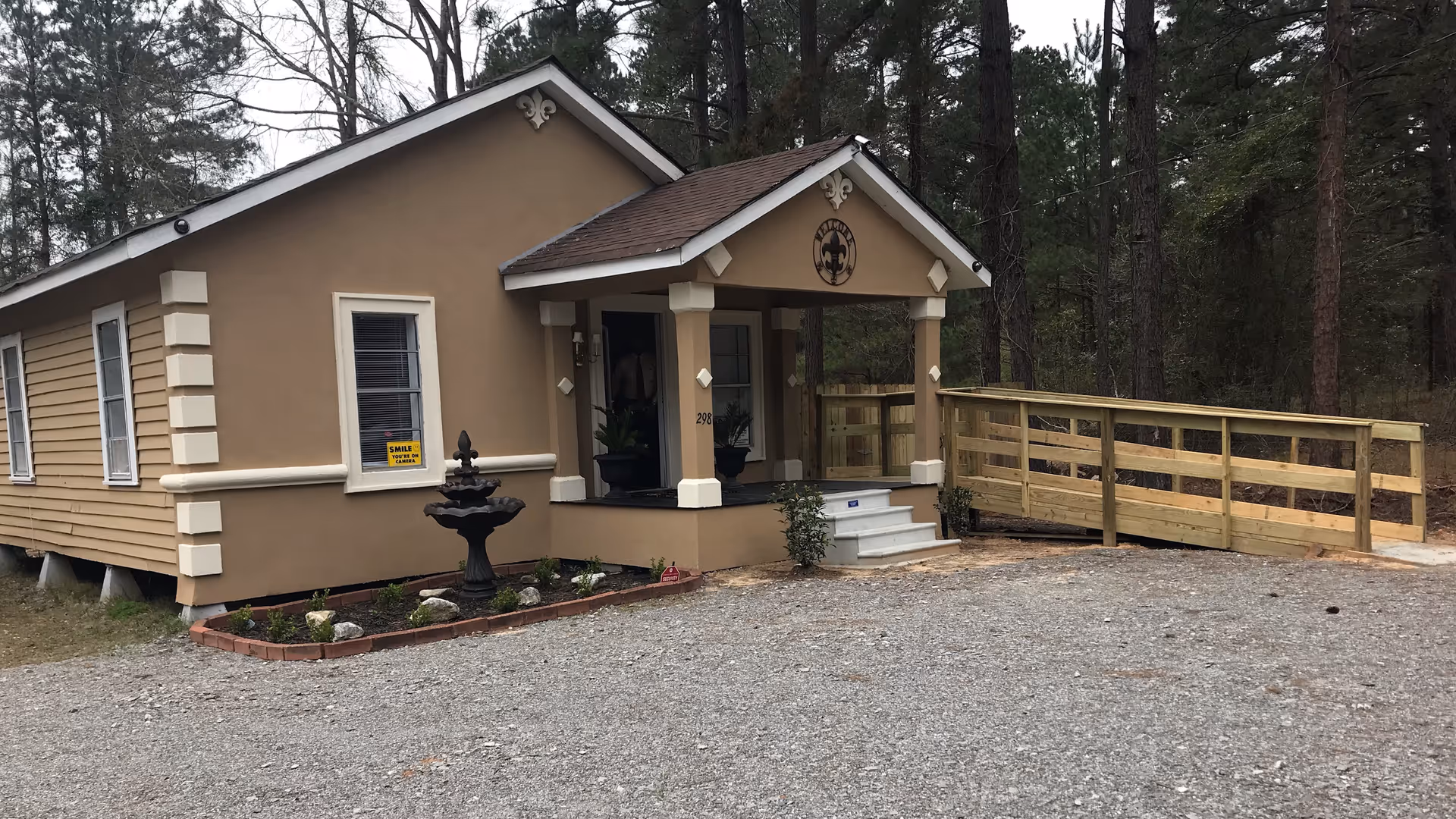 Small beige hospice building with front porch, steps, and a wooden ramp surrounded by trees.