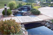 Water feature with a small waterfall, stone steps, and a wooden footbridge in a landscaped area.
