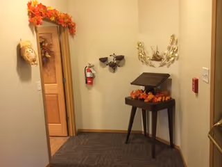 A small hallway corner decorated with autumn-themed decorations including a garland of orange leaves above a doorway, a small table with fall leaves and a decorative birdhouse, and wall-mounted metal bird sculptures. A fire extinguisher is mounted on the wall, and the floor is carpeted.