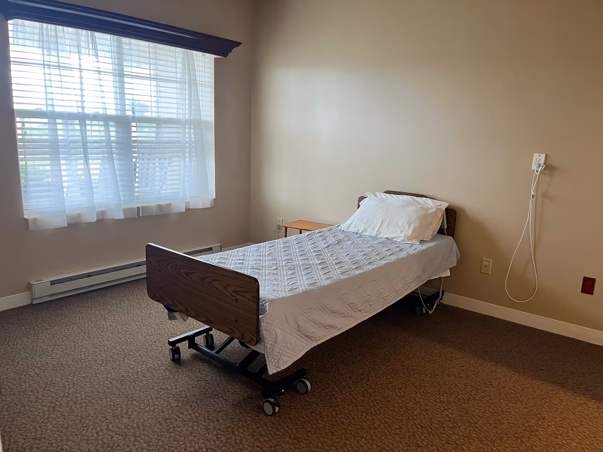 A simple bedroom in a senior care facility with a single hospital-style bed covered with a white quilt and pillow. The room has beige walls, a large window with sheer white curtains, a small wooden side table, and a call button with a cord on the wall.