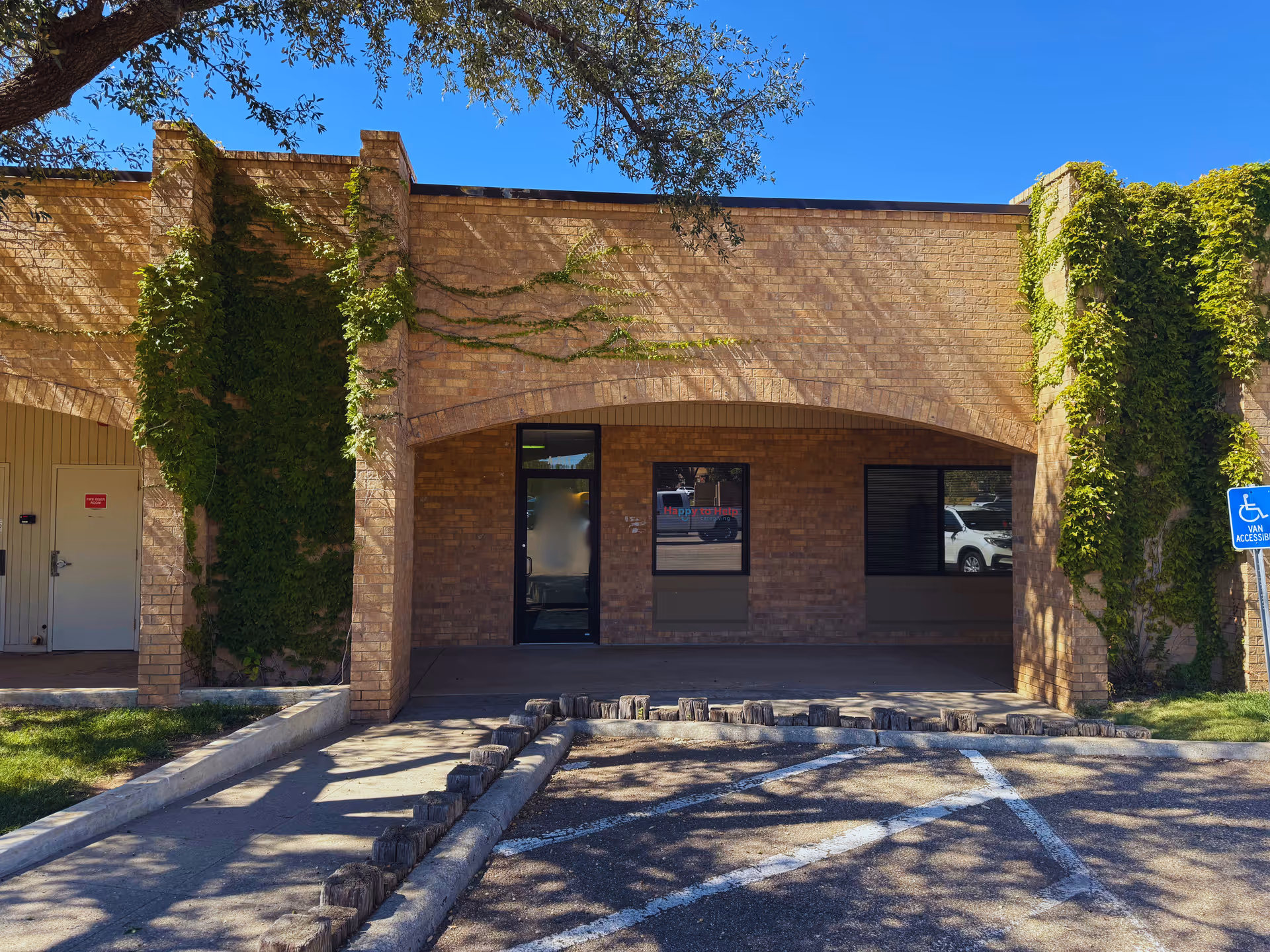 Brick building with ivy-covered walls, arched entrance, windows, and a parking lot under blue sky.