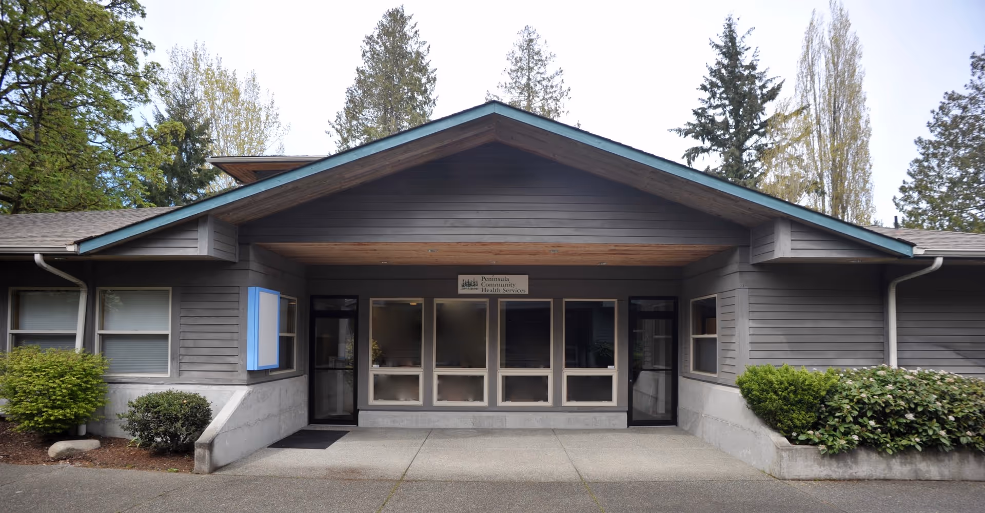 Gray single-story clinic with glass doors and surrounding shrubs at the entrance.