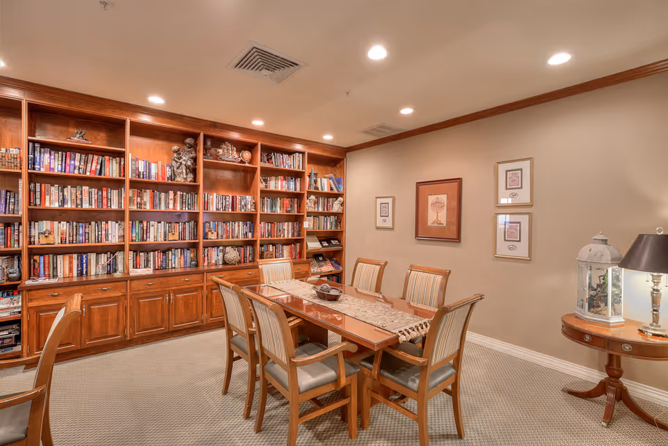 A cozy room with a large wooden bookshelf filled with books along one wall, a wooden table with six chairs in the center, and a small round side table with a lamp and decorative items against the opposite wall. The room has beige walls, carpeted floor, and recessed ceiling lights.