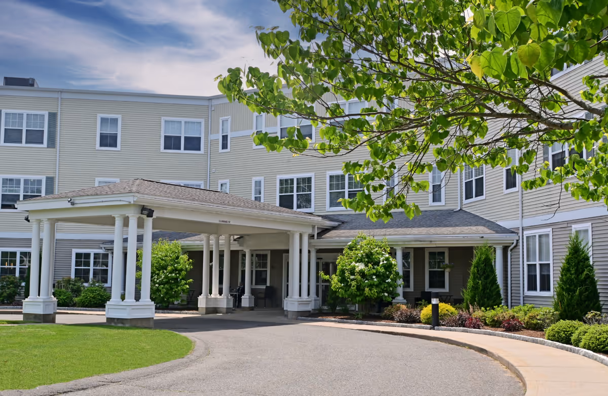 Front entrance of a multi-story residential building with a covered porte-cochere, driveway, and landscaped grounds.