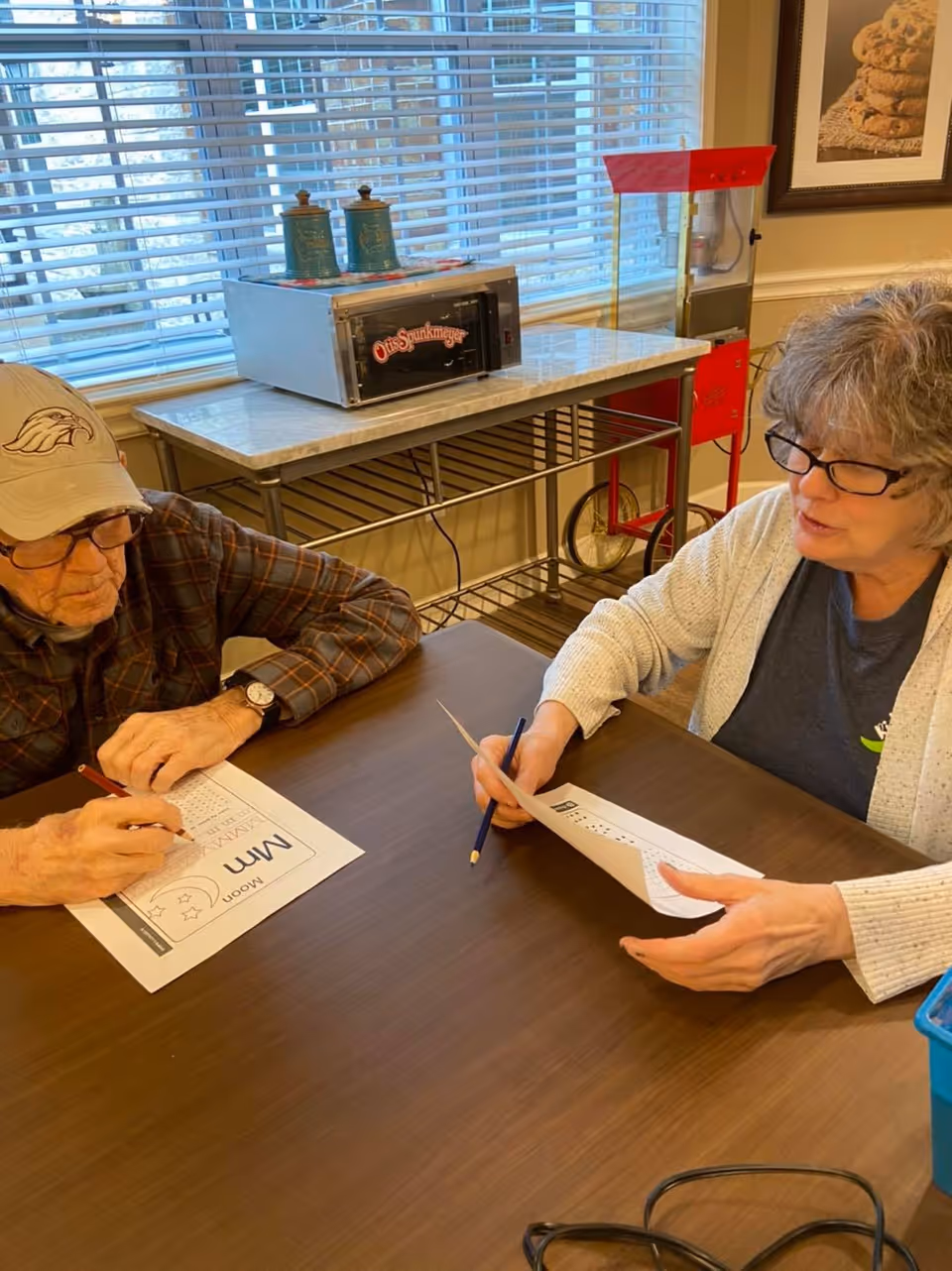 An elderly man and a woman sitting at a table engaged in an activity with printed worksheets and pencils. The man is wearing a beige cap and glasses, while the woman has short gray hair and glasses. Behind them is a window with blinds, a table with a vintage popcorn machine, and a framed picture on the wall.