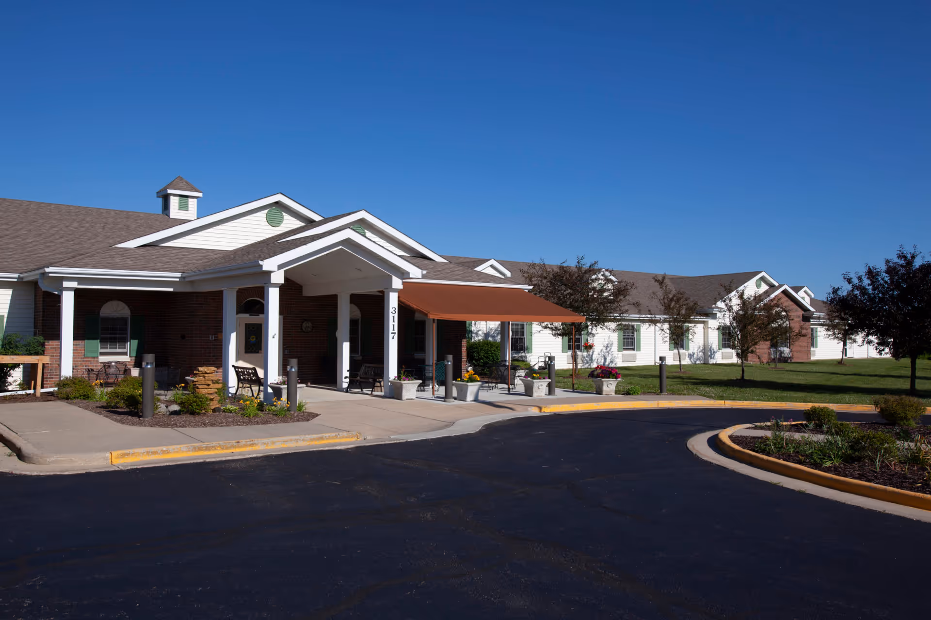 Exterior view of Oakley Courts Assisted Living Community building with a covered entrance, benches, landscaped garden beds, and a clear blue sky.