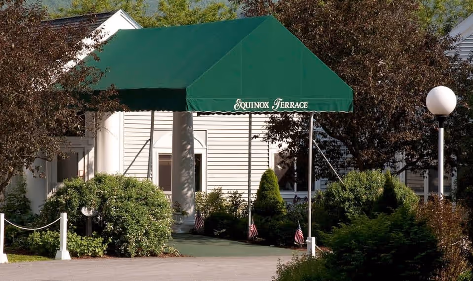 Entrance to a building with a green canopy that reads 'Equinox Terrace'. The building is white with columns and surrounded by bushes and trees. There is a white lamp post on the right side and a paved driveway leading to the entrance.