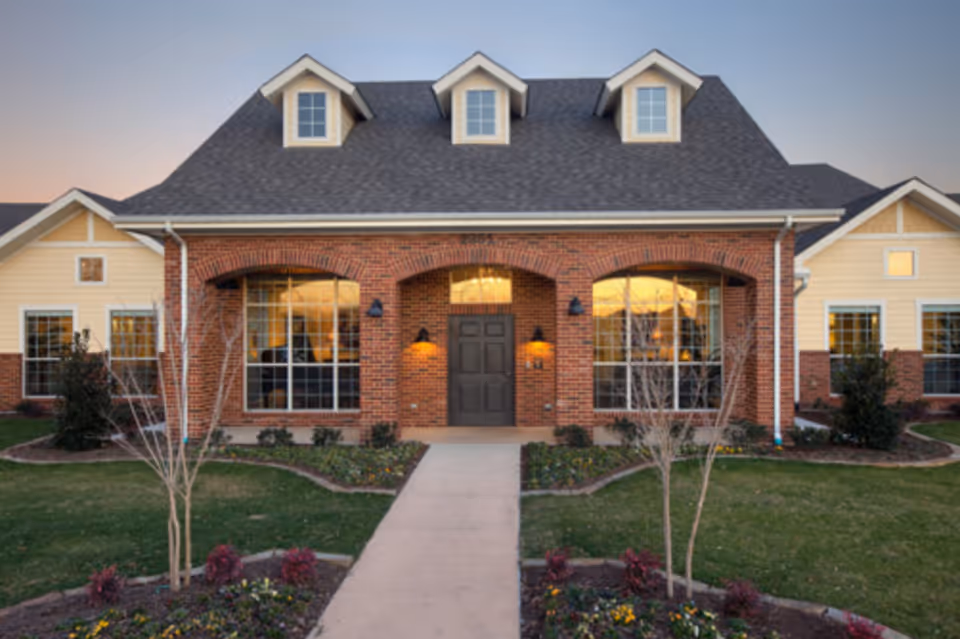 Front entrance of a single-story brick-faced cottage with large windows, dormer windows, and a landscaped walkway.