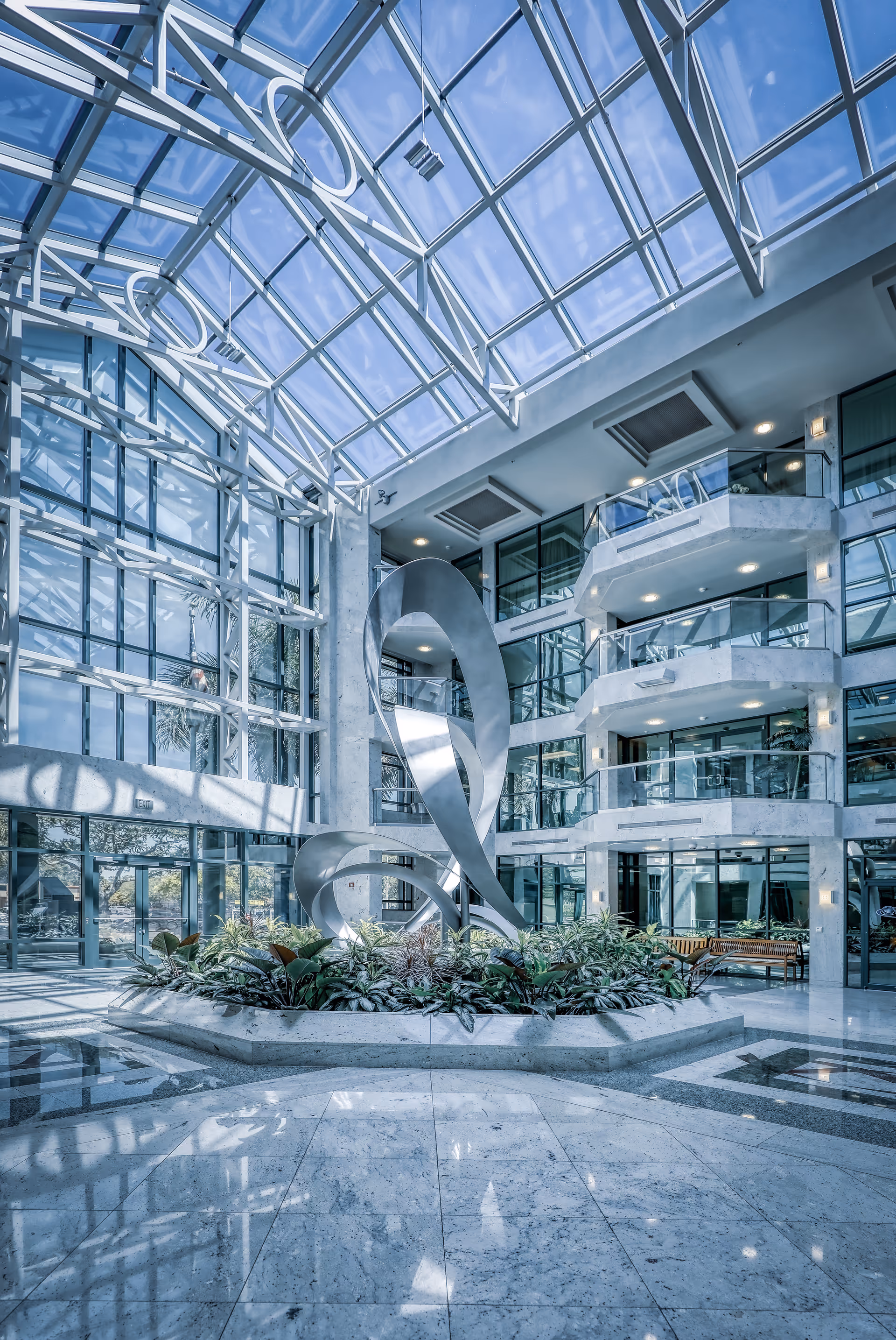 Bright glass atrium with a sculptural centerpiece, indoor plants, and balconies.