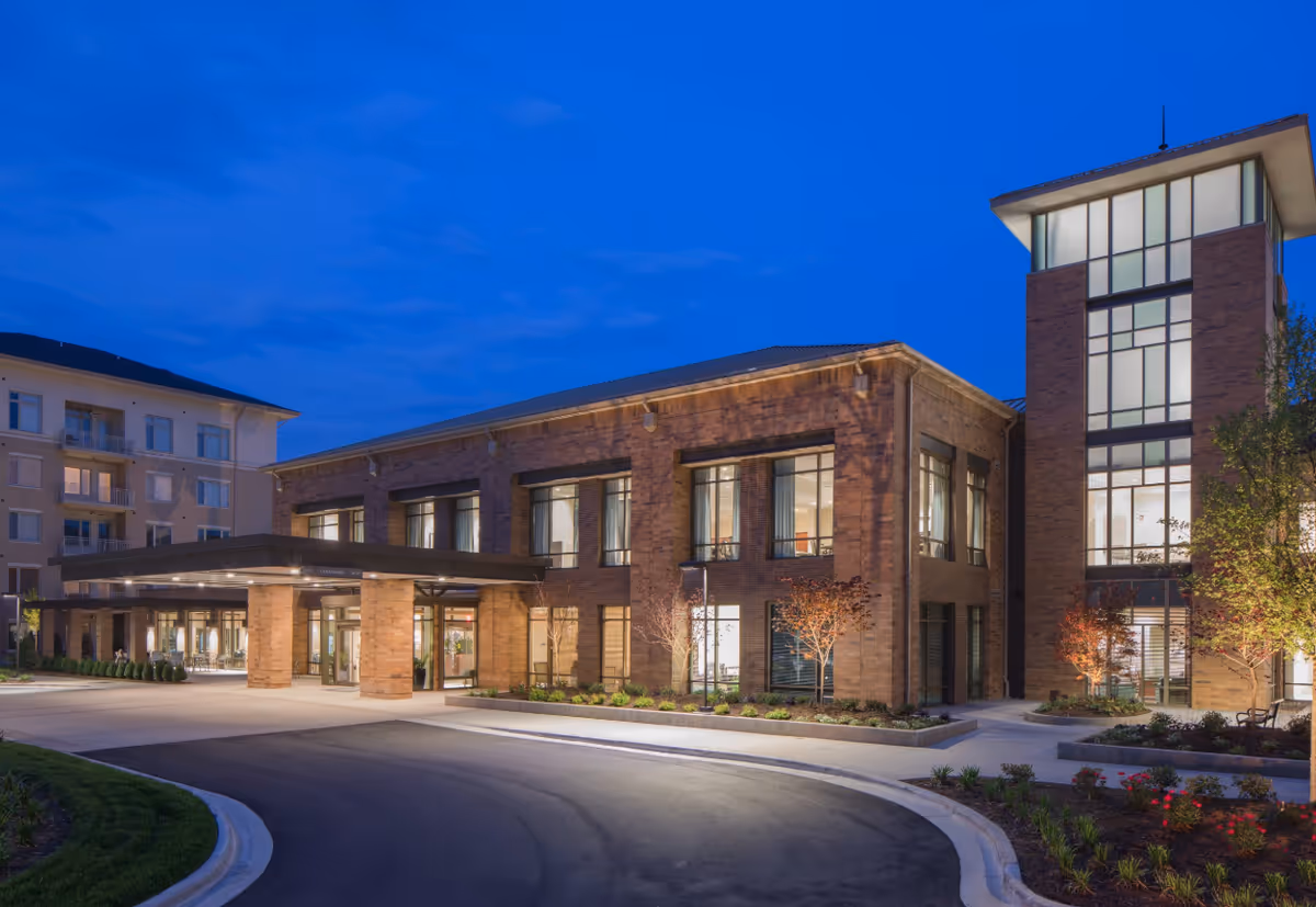 Exterior view of a modern senior living facility building at dusk with large windows, brick facade, and a covered entrance driveway surrounded by landscaped greenery.