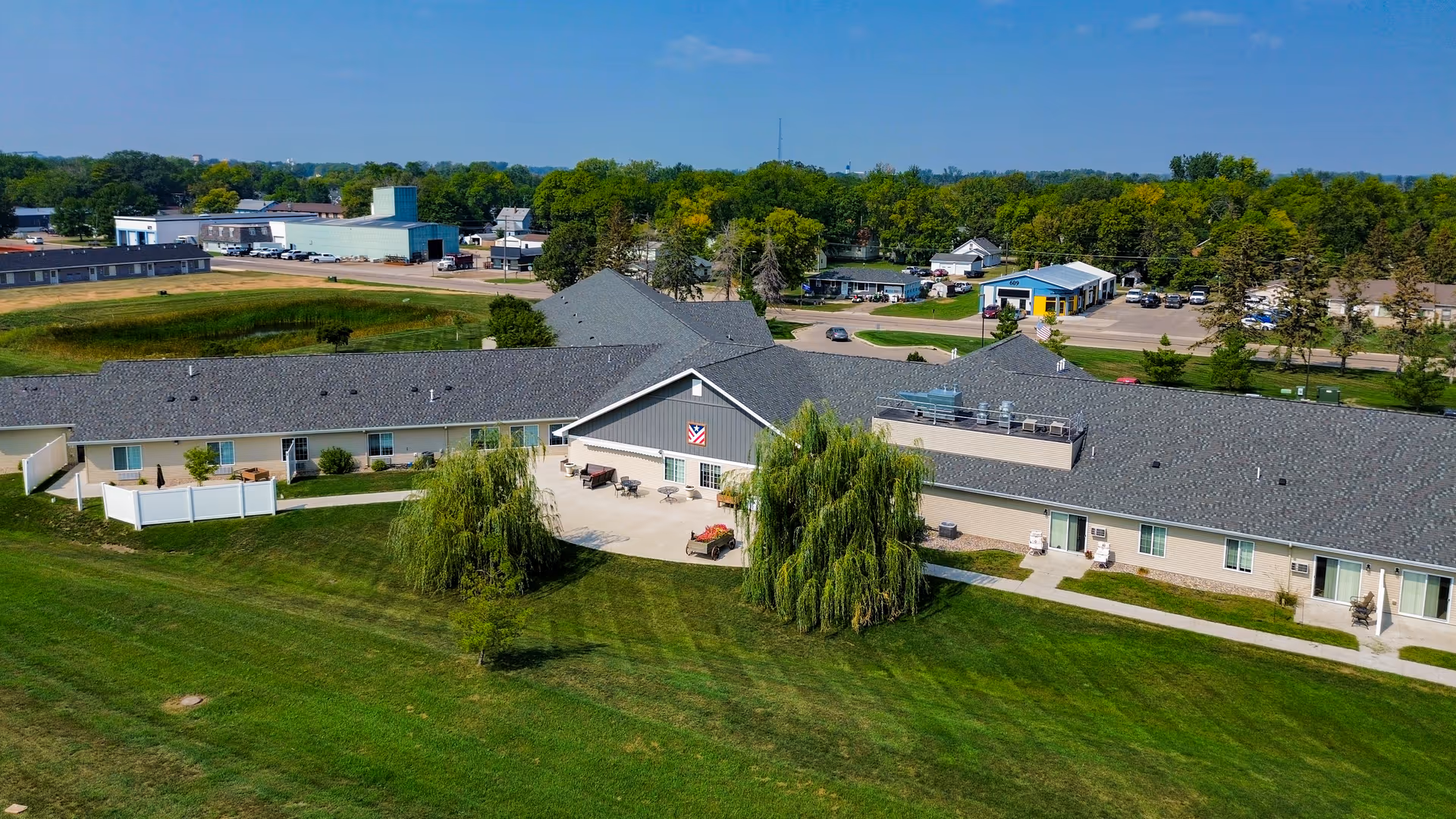 Aerial view of The Legacy Assisted Living facility in Morris, MN, showing a large single-story building with a gray roof surrounded by green lawns and trees. There is a central courtyard area with outdoor seating and a few small trees. The background includes other buildings, parking areas, and a clear blue sky.