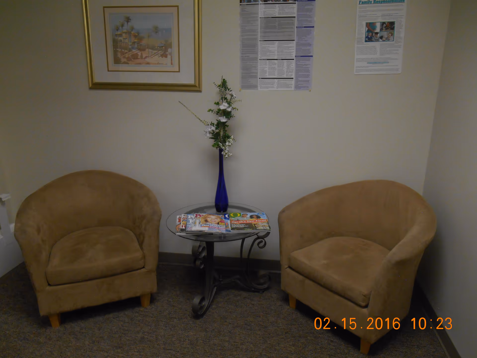 Two beige armchairs flank a glass table with magazines and a tall blue vase.