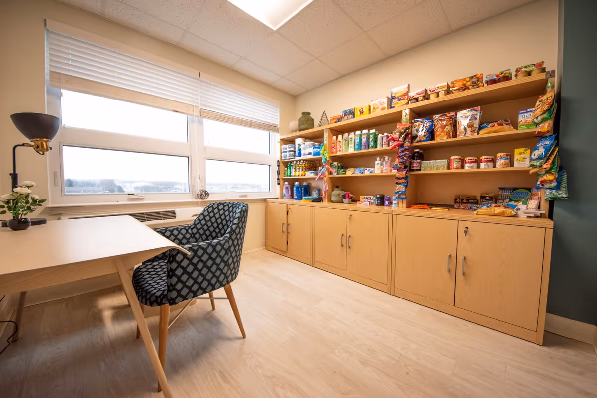 A bright interior room with a table and patterned chair by a large window and shelves stocked with snacks and household items.