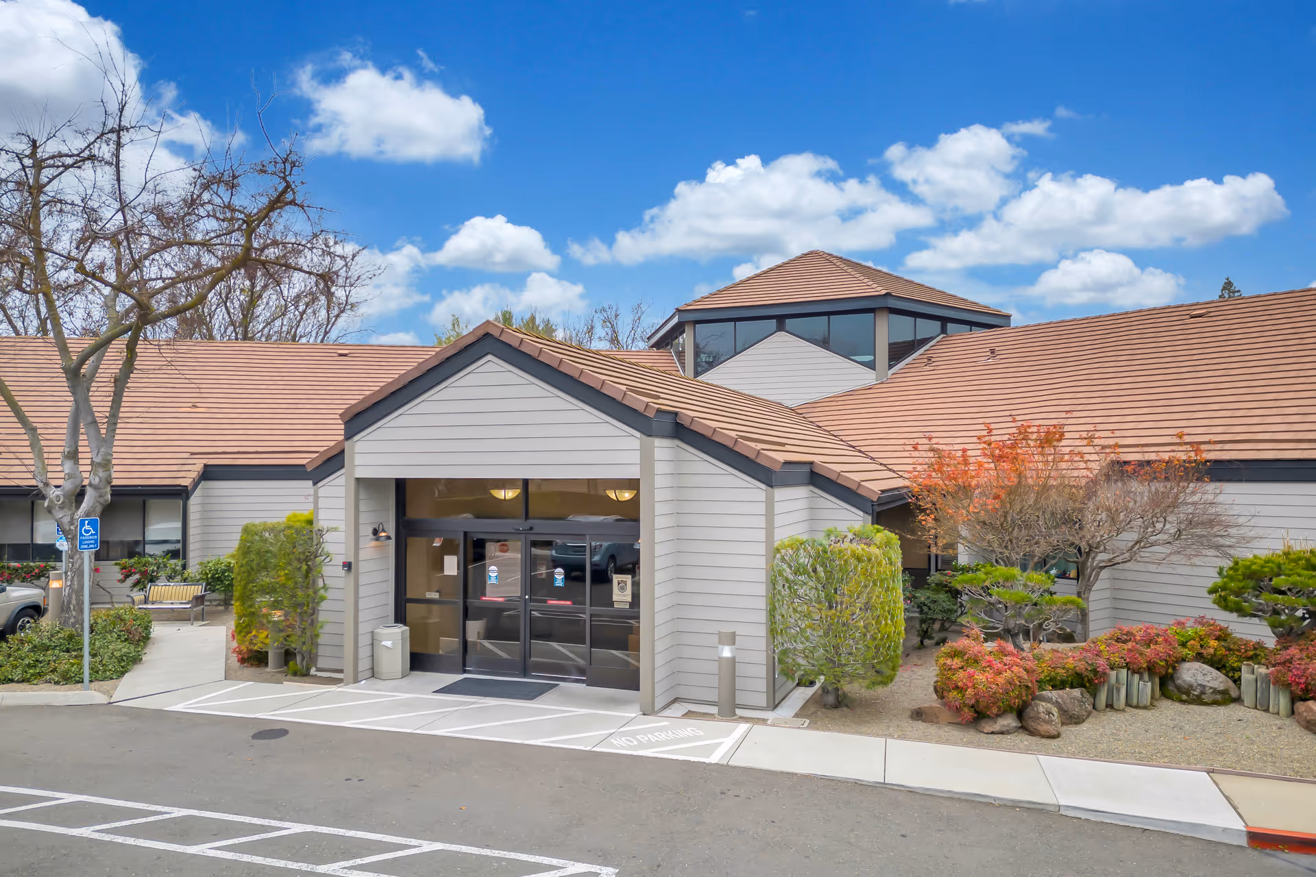 Exterior view of ACC Care Center: Skilled Nursing & Rehabilitation building entrance with a brown tiled roof, beige siding, glass double doors, landscaped bushes, trees, and a clear blue sky with clouds.