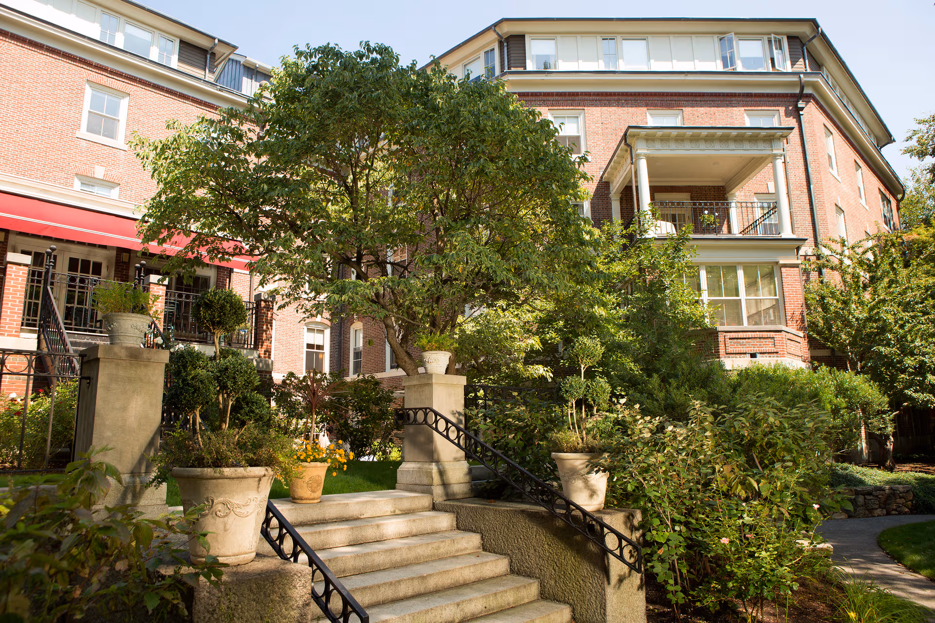 Brick senior living building with stairs, lush greenery, and a red awning over the entrance.
