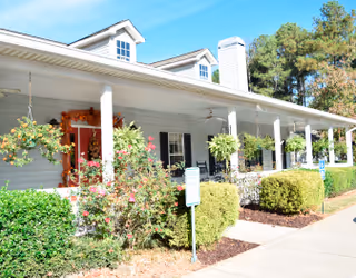 Front porch of a single-story assisted living building with white columns, hanging plants, shrubs, and a paved walkway.