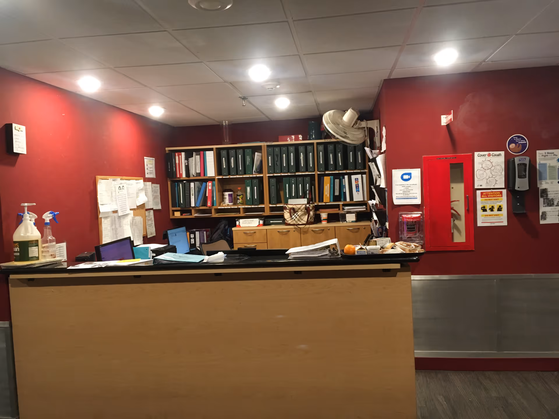Reception desk in a red-walled interior, with shelves, paperwork, and cleaning supplies.