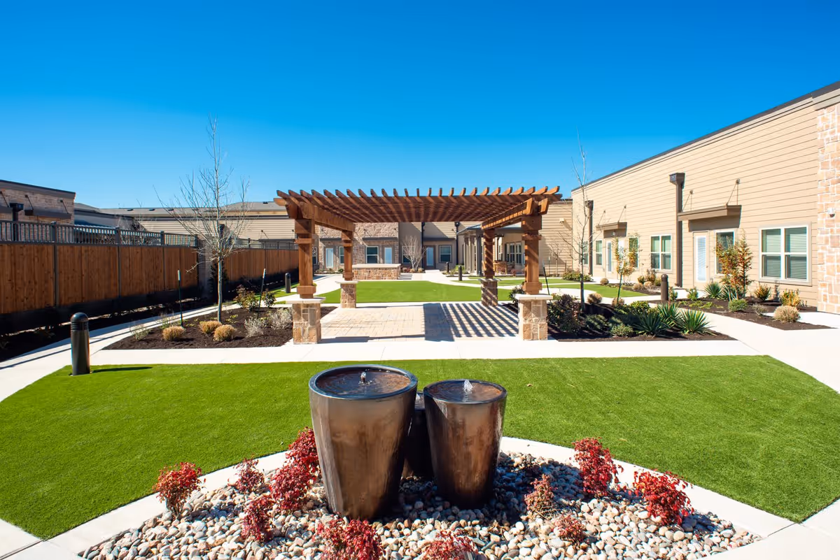 Outdoor courtyard area at Discovery Village at Alliance Town Center Assisted Living featuring a wooden pergola, manicured green lawns, a stone pathway, decorative plants, and two modern water fountains in the foreground under a clear blue sky.
