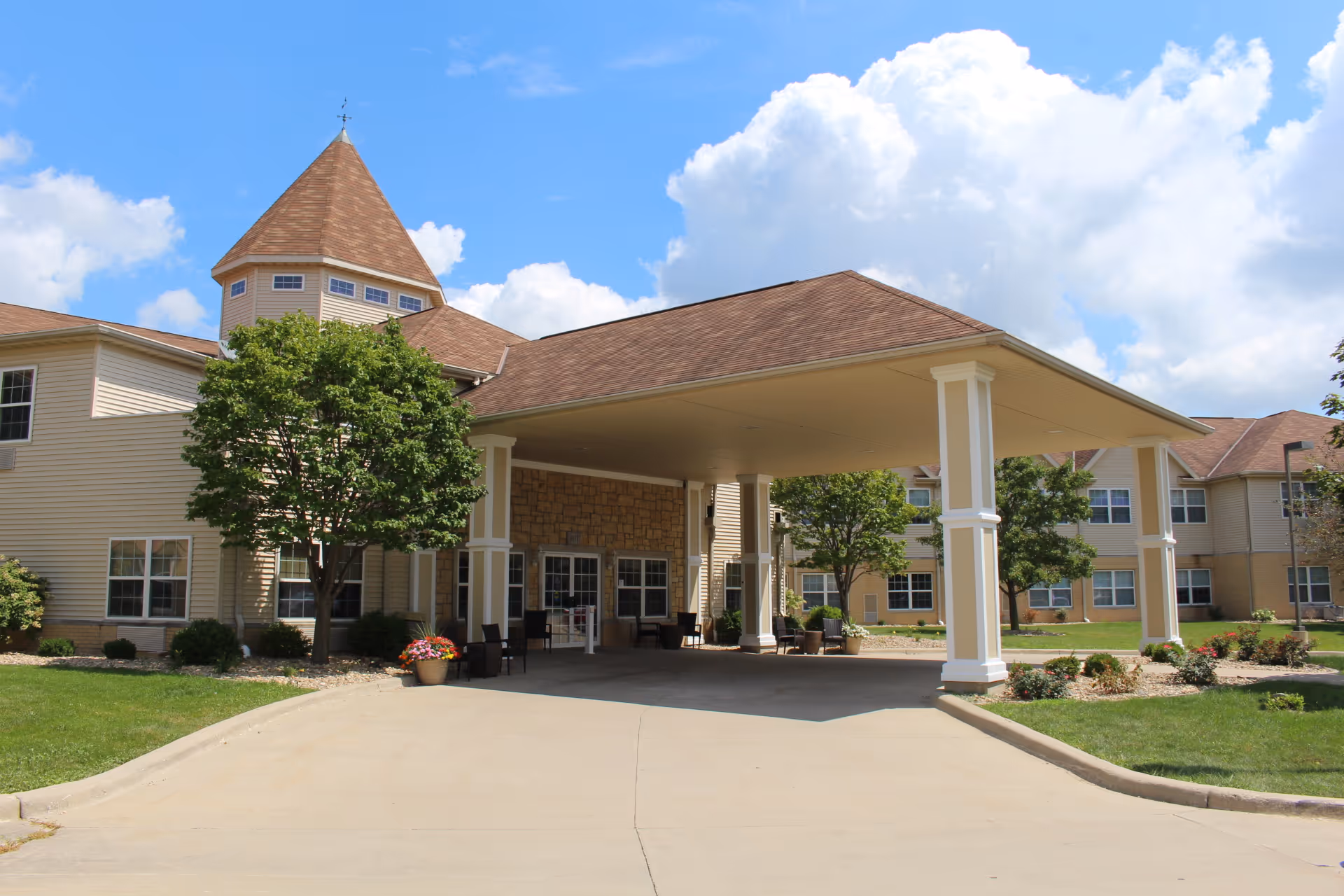 Exterior view of a senior living facility with a covered entrance supported by white columns, beige siding, and a conical roof tower. There are trees, shrubs, and flower pots around the entrance, with a clear blue sky and some clouds in the background.
