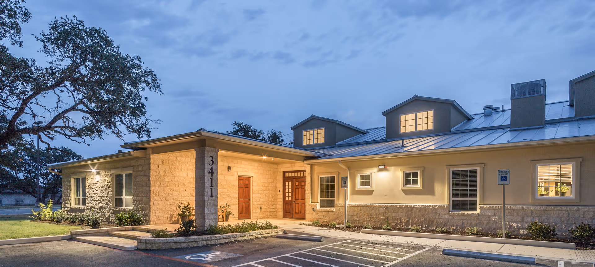 Exterior view of a single-story building at dusk with warm lights glowing from the windows. The building has a stone and stucco facade with a metal roof and two dormer windows. There is a parking area in front with designated handicap parking spaces and a sidewalk leading to a wooden double door entrance under a covered porch. Trees and landscaping surround the building.