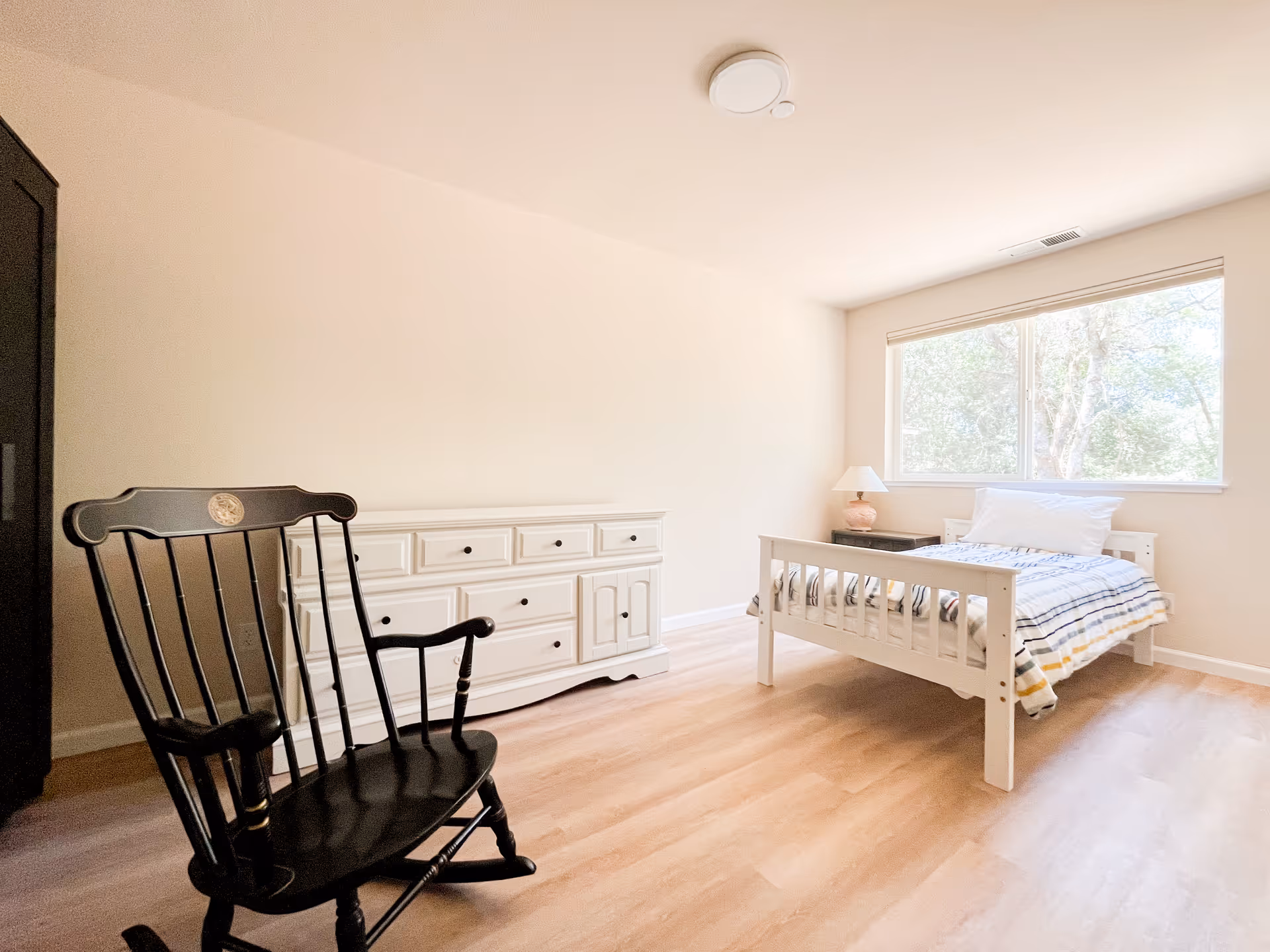 Bright senior living facility bedroom with a white dresser, bed, nightstand, and rocking chair.