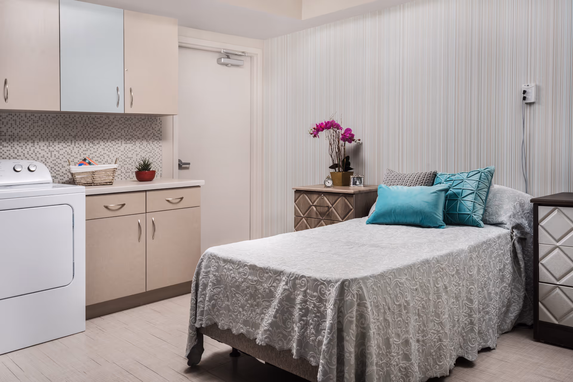 A compact bedroom with a bed, pillows, nightstand, and a kitchenette in a senior facility.