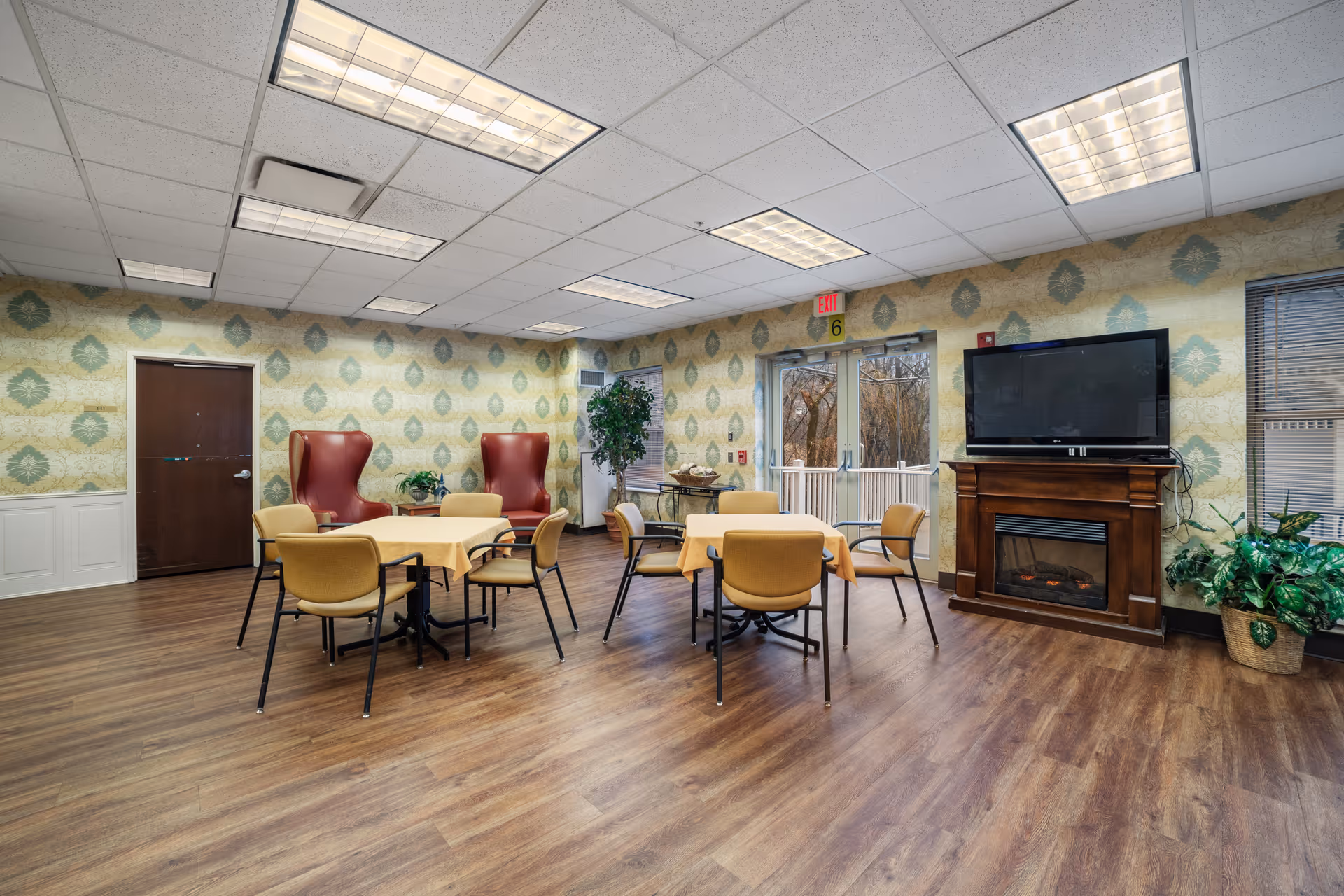 Senior living common room with round tables, chairs, wallpaper, and a fireplace with TV