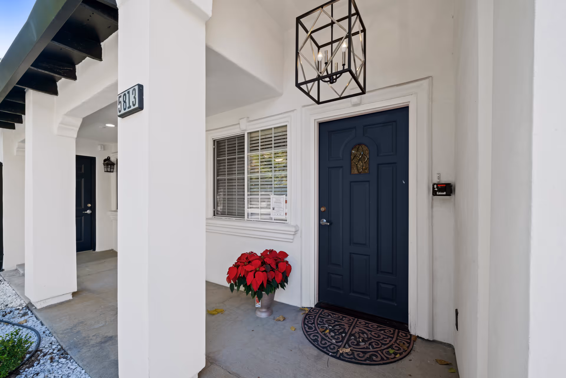 Entrance to a senior living facility unit with a dark blue door, a decorative doormat, a potted plant with red flowers, white walls, and a hanging lantern-style light fixture. The unit number 5813 is displayed on a white pillar near the door.