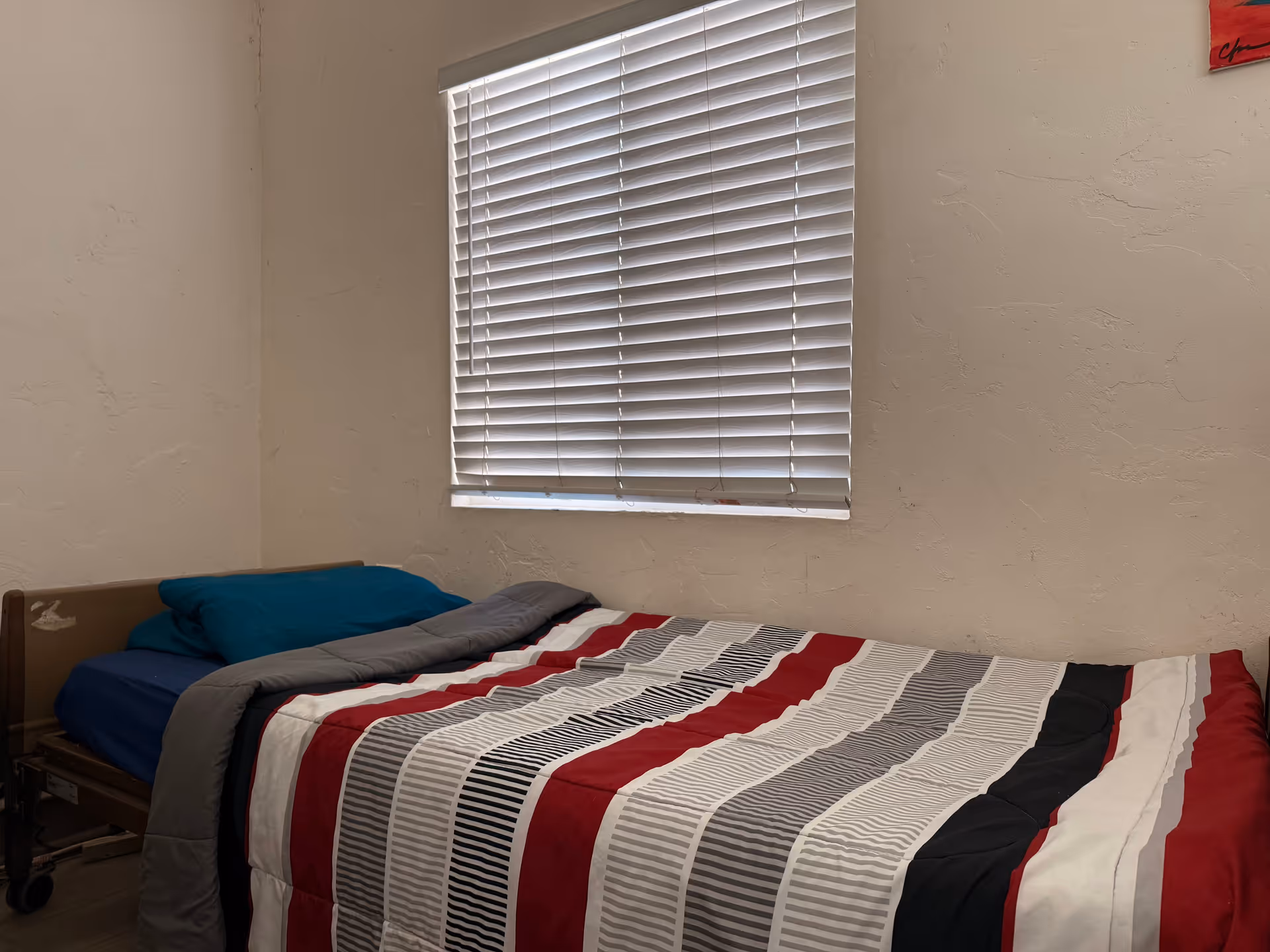 A bedroom with a striped bedspread, blue pillow, and window blinds on plain walls.