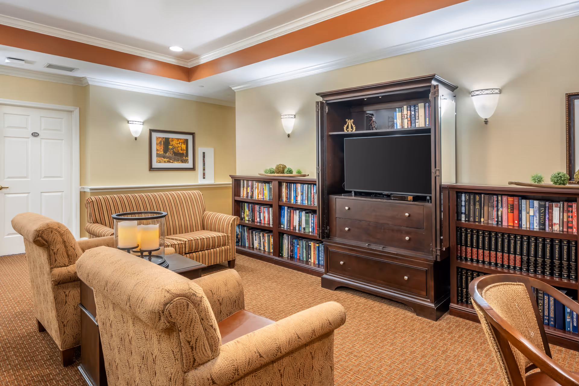A cozy living room area with two upholstered armchairs and a striped loveseat arranged around a small wooden coffee table with three candles. There is a large dark wooden entertainment center with a TV and bookshelves filled with books on either side. The walls are beige with framed artwork and wall sconces providing soft lighting. The carpet is a warm brown color, and there is a white door labeled 220 in the background.