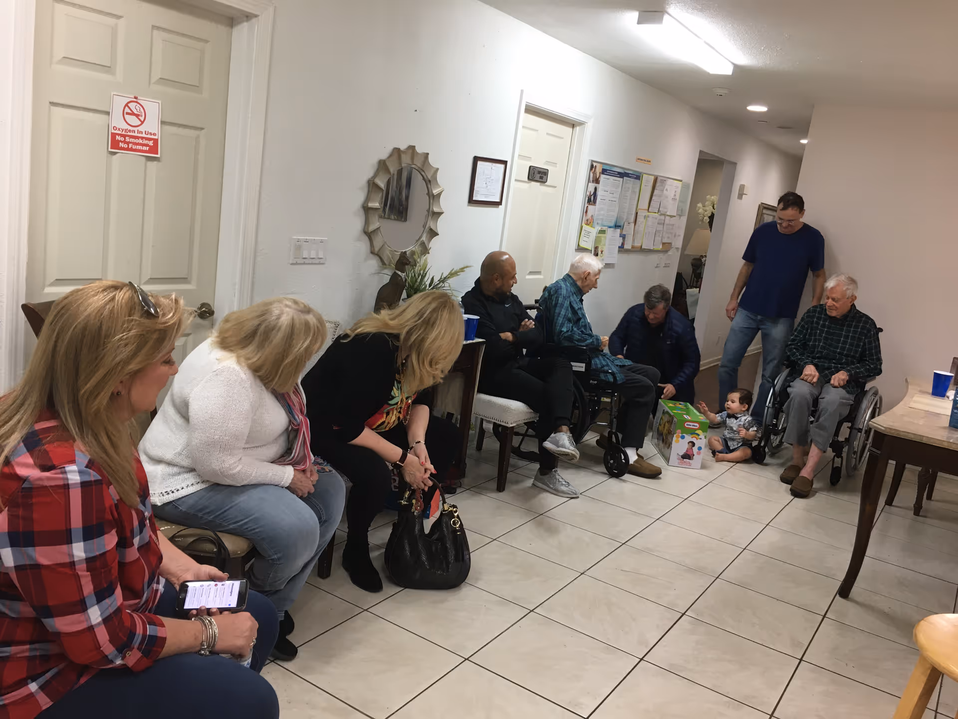 A group of people, including elderly individuals and adults, are seated and interacting in a hallway or common area of a care home. Some elderly men are in wheelchairs, and a young child is sitting on the floor playing with a toy box. The walls are white with a decorative mirror and bulletin board visible. The floor is tiled, and the lighting is bright with ceiling lights.