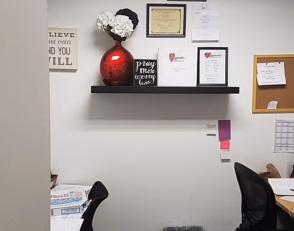 Office corner with shelf, framed documents, red vase, bulletin board, chairs, and desk.