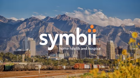 City skyline with mountains in the distance; foreground shows railroad tracks and yellow flowers.