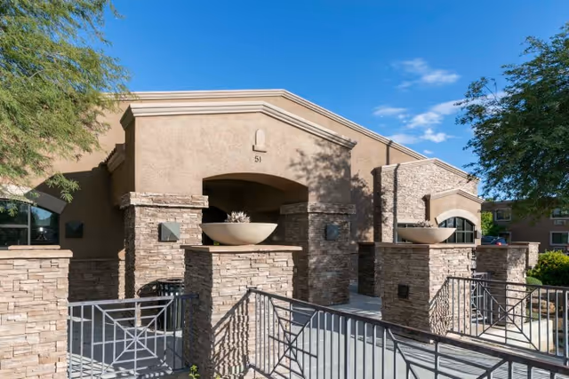 Exterior view of a building with stone pillars and beige stucco walls under a clear blue sky. The entrance features large planters on stone pedestals and metal railings leading up to the doorway. Trees and greenery surround the area.