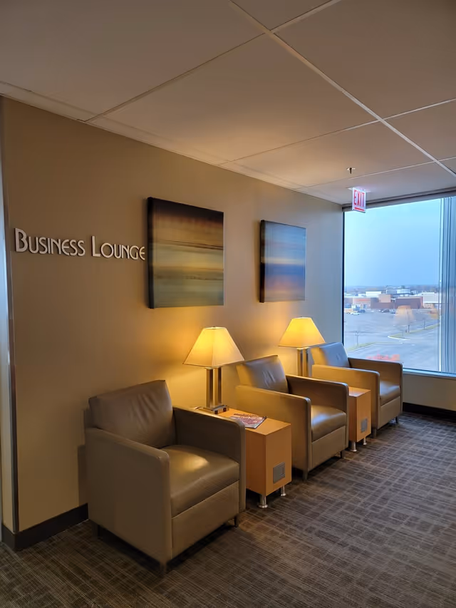 Four beige armchairs with side tables and lamps in a business lounge by a window.