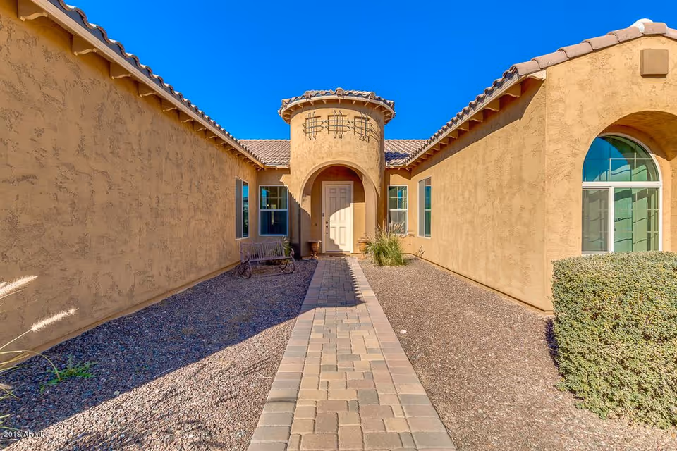 Paved walkway leading to an arched entryway of a tan stucco building with a tiled roof under a clear blue sky.