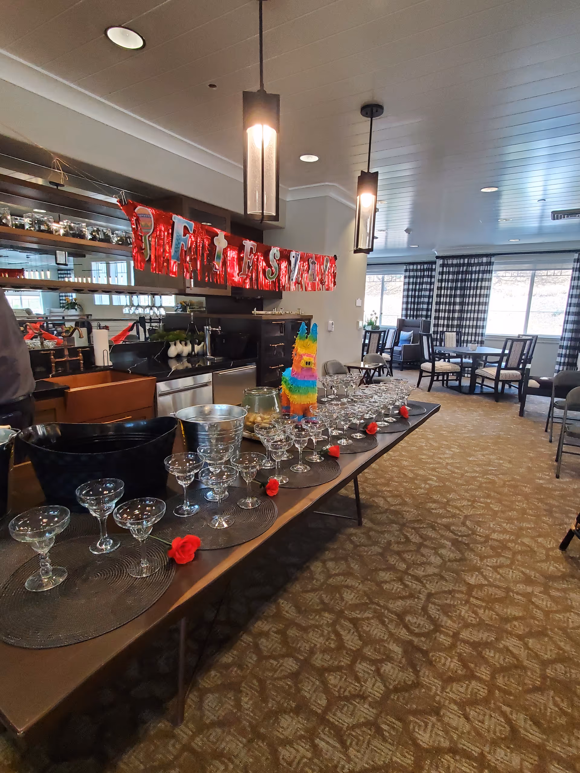 Long dining table with numerous wine glasses and birthday banner inside a care facility dining room.
