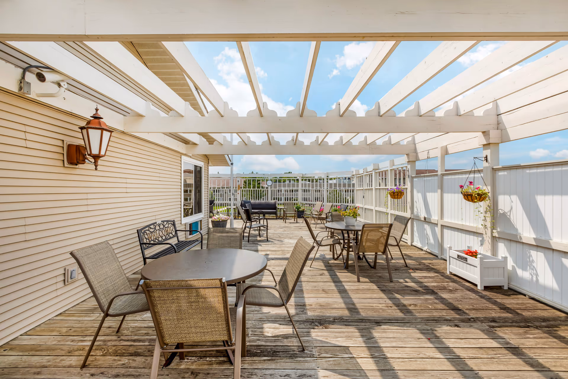 Outdoor wooden deck with tables and chairs under a white pergola, potted plants along railing.