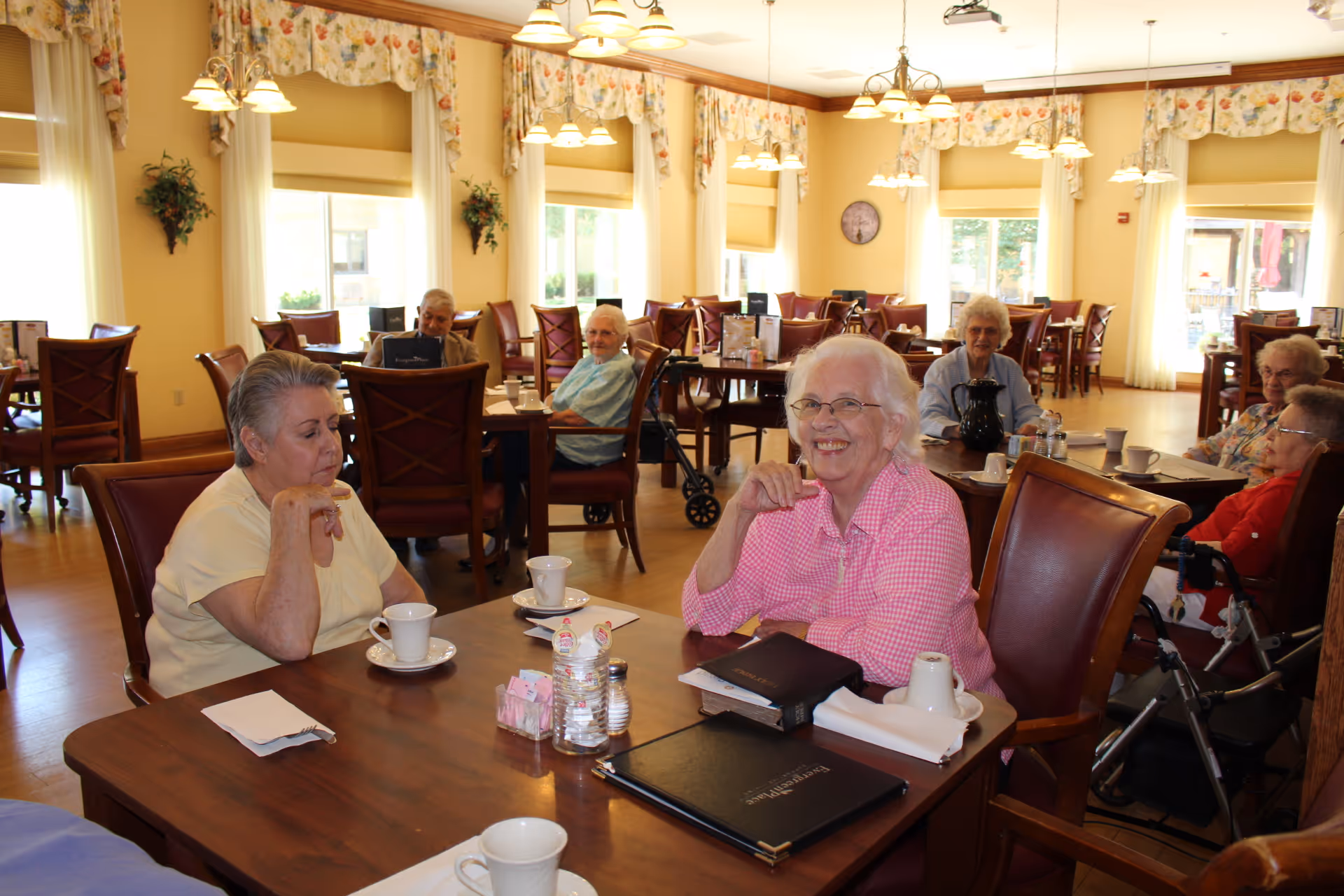 A group of elderly women sitting at tables in a bright dining room with large windows, floral curtains, and warm yellow walls. They are enjoying drinks and conversation, with menus and cups on the tables.