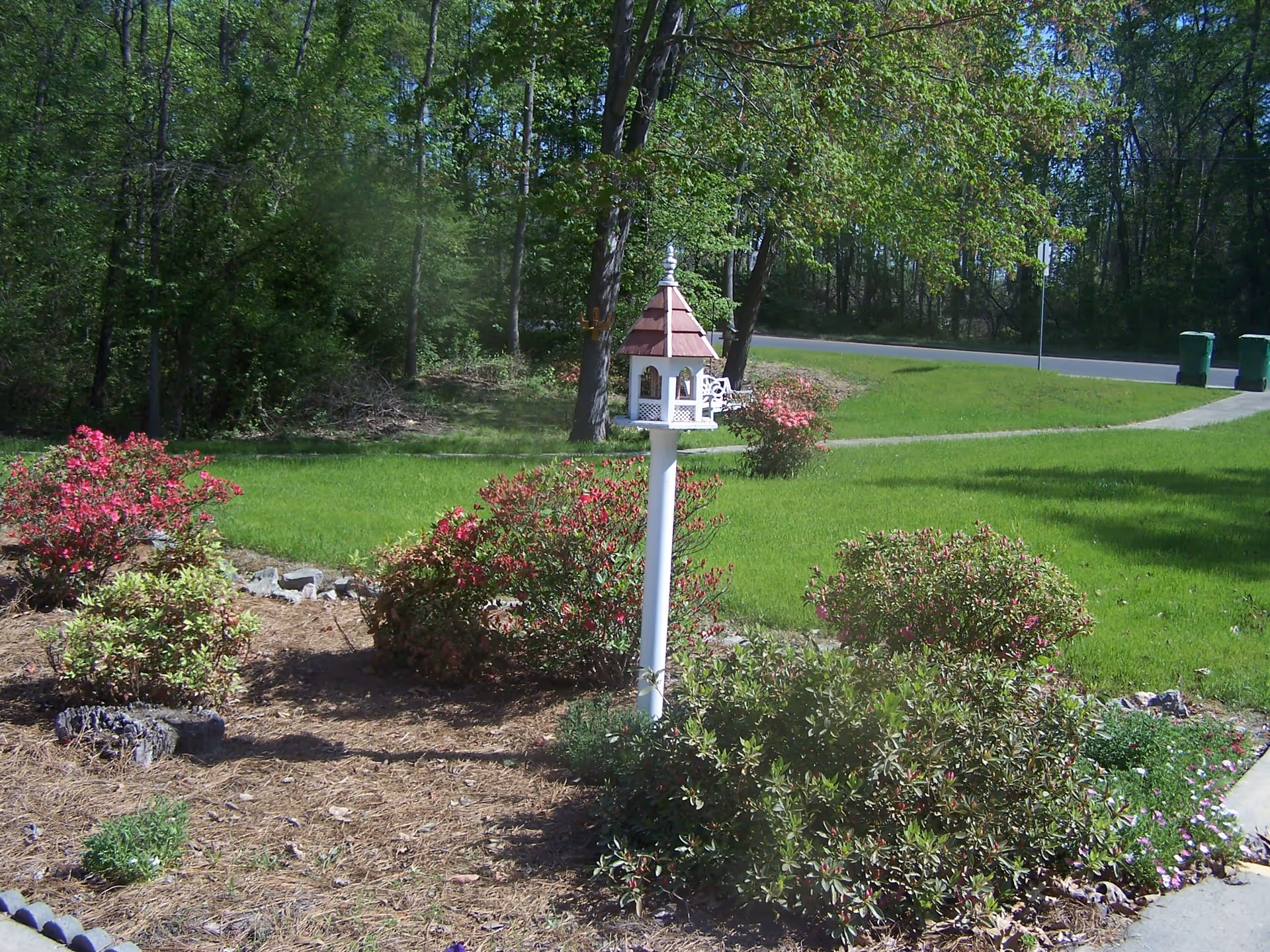 A landscaped front yard with shrubs and a decorative birdhouse on a white post beside a driveway and road.