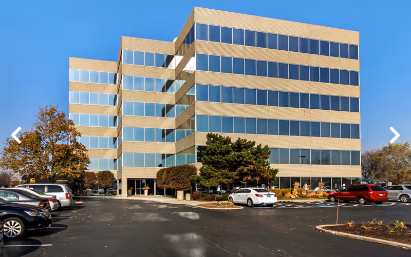 A modern brick-and-glass office building with multiple stories and a parking lot in front.