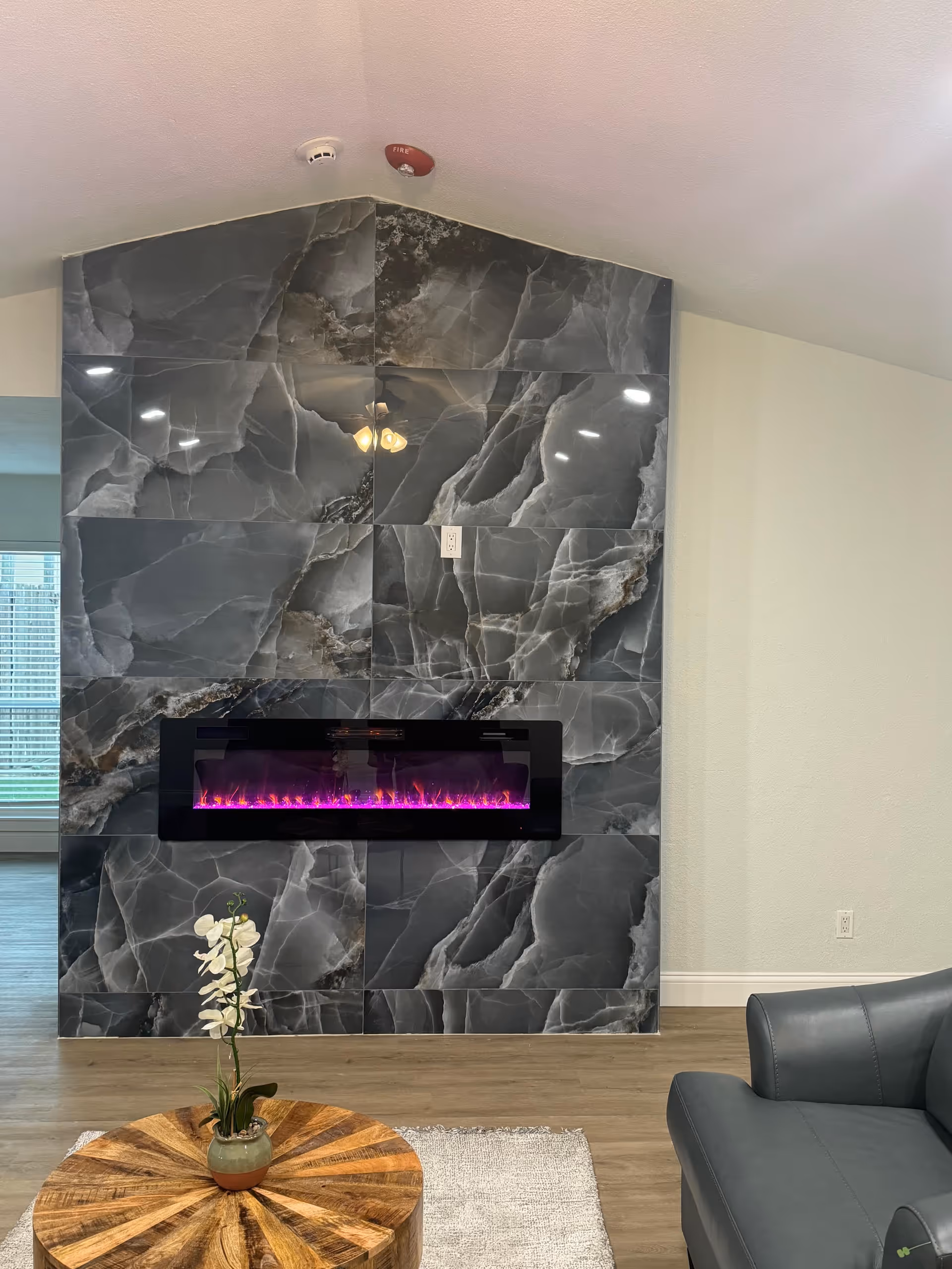 Modern living room features a marble-tile wall with a purple electric fireplace and a round table.