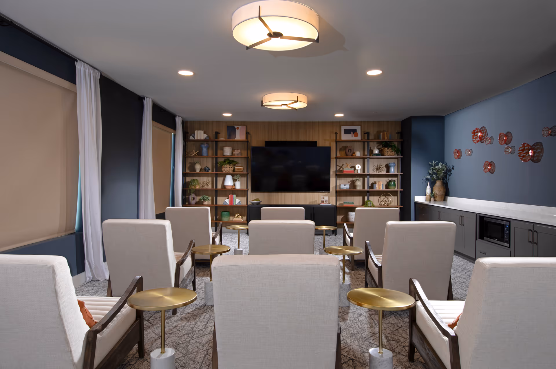 A modern common room with beige armchairs arranged in rows facing a large flat-screen TV mounted on a wooden paneled wall. The room features built-in shelves with decorative items, a long countertop with cabinets and a microwave, blue walls with red wall art, and two ceiling lights.
