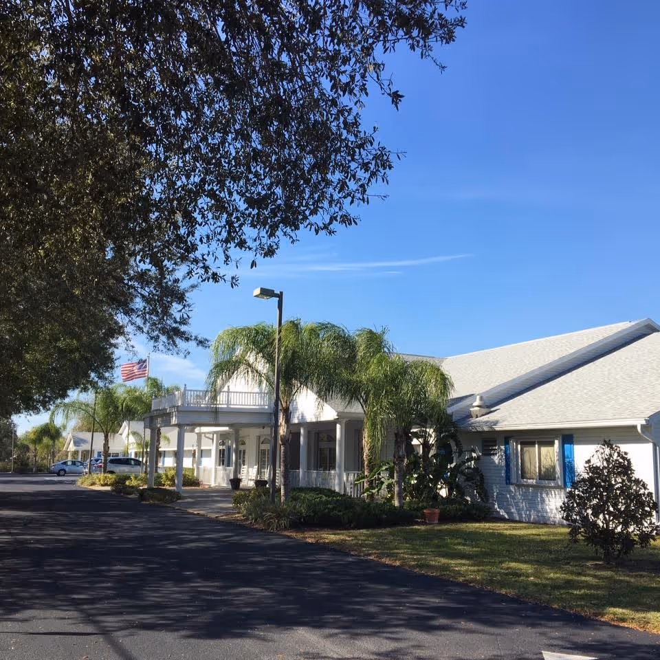 Exterior view of a single-story building with white walls and a light gray roof, surrounded by palm trees and other greenery under a clear blue sky. There is a paved driveway or parking area in front, and an American flag is visible in the background.