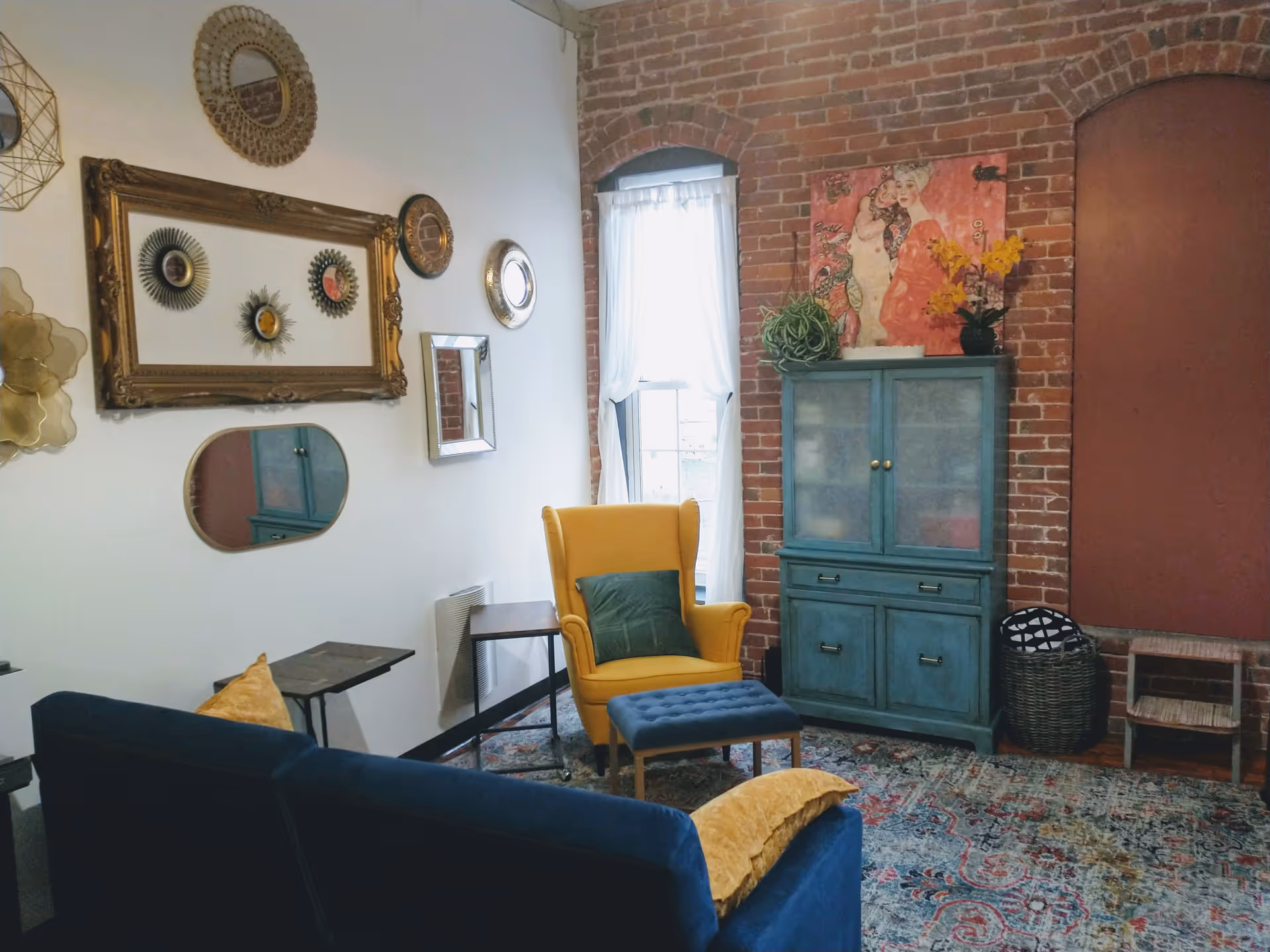 A cozy living room with a yellow chair, blue cabinet, brick wall, and colorful decor.