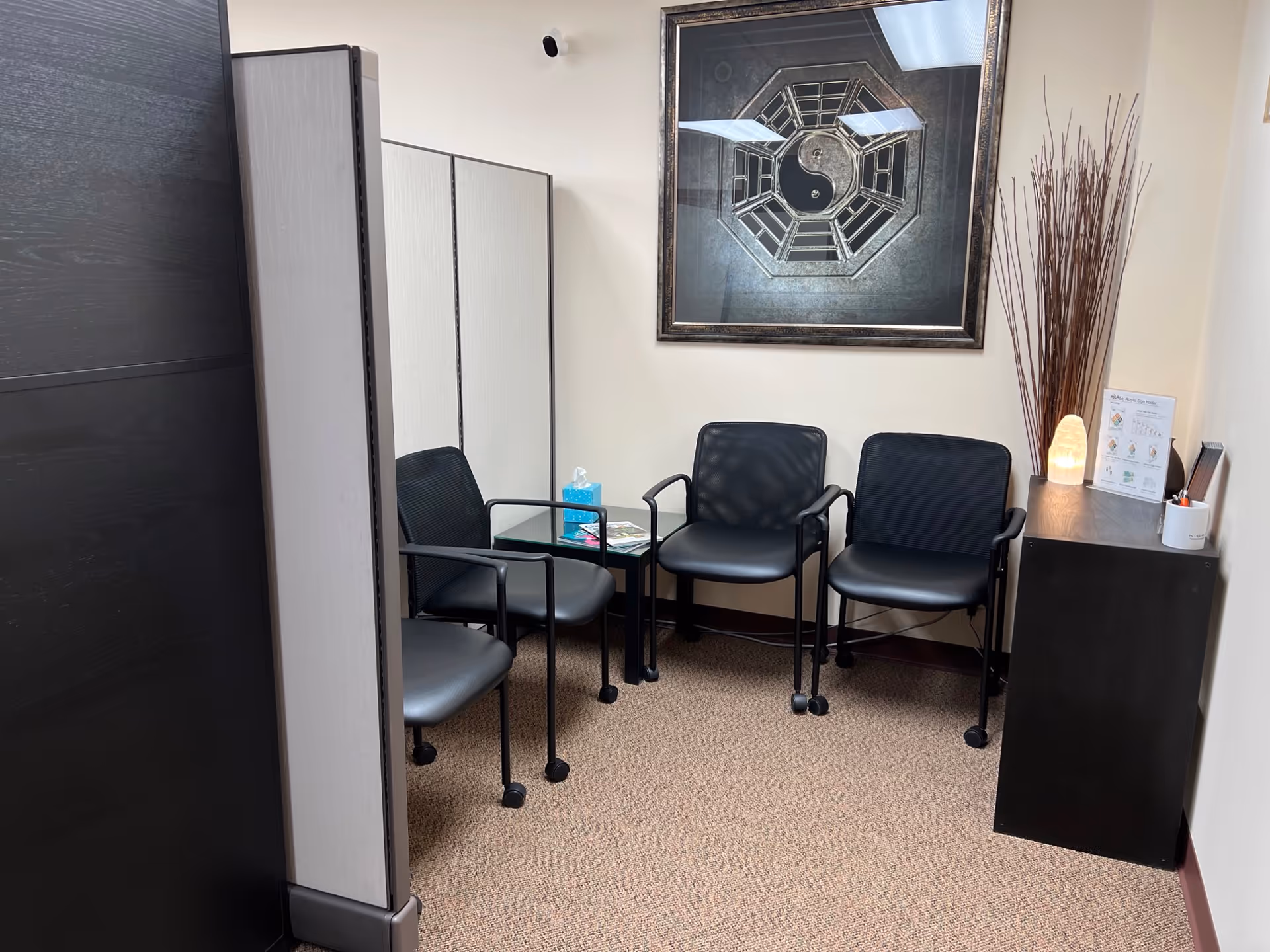 A small waiting area with black chairs, partition, and decorative wall art in a beige hallway.